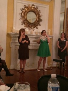 Kathy MacMillan stands in front of a mantelpiece speaking into a microphone while a woman beside her interprets in American Sign Language.