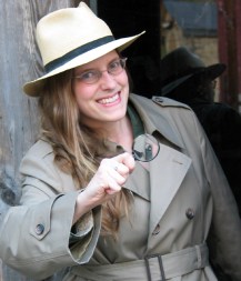 photo photo of Kathy MacMillan in an overcoat and fedora, holding a magnifying glass