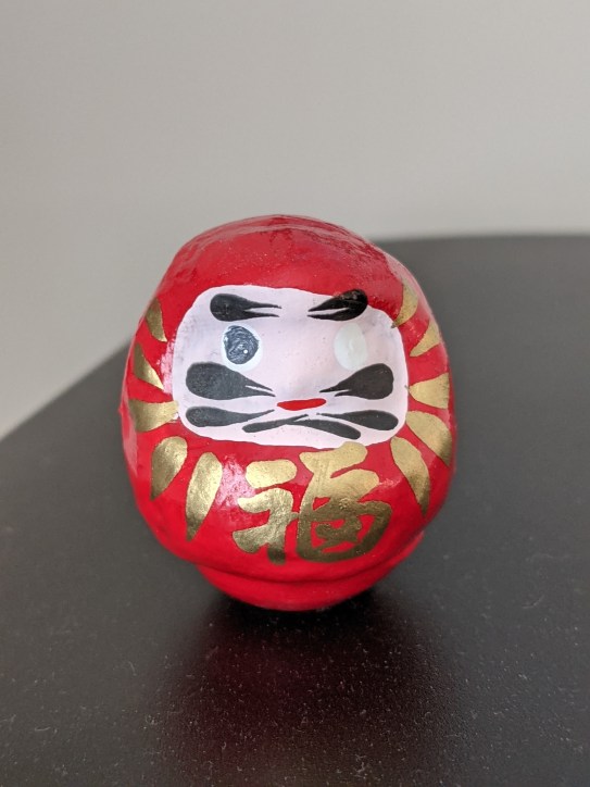 a round red daruma doll sits on a desk. It has a pale face with black markings. Golden symbols are painted around the face.