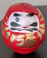 a round red daruma doll sits on a desk. It has a pale face with black markings. Golden symbols are painted around the face.