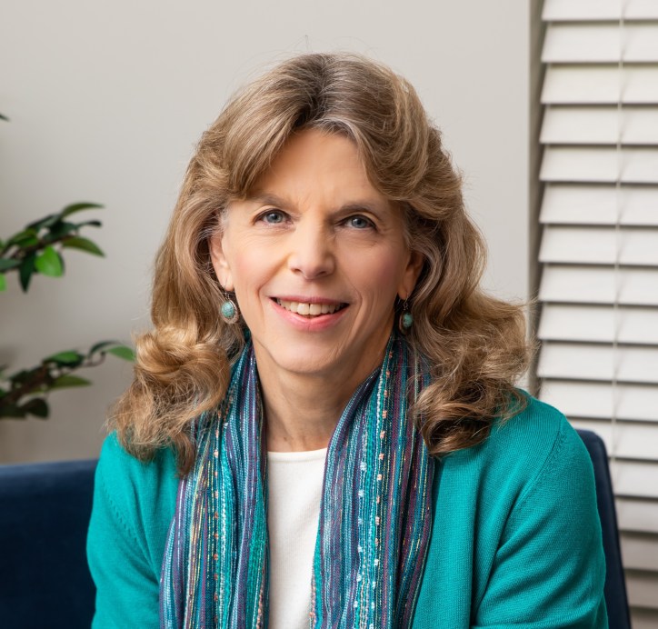 A white woman with light brown hair smiles at the camera. She is wearing a turquise jacket over a white shirt. A patterned turqoise scarf hangs loose around her neck.